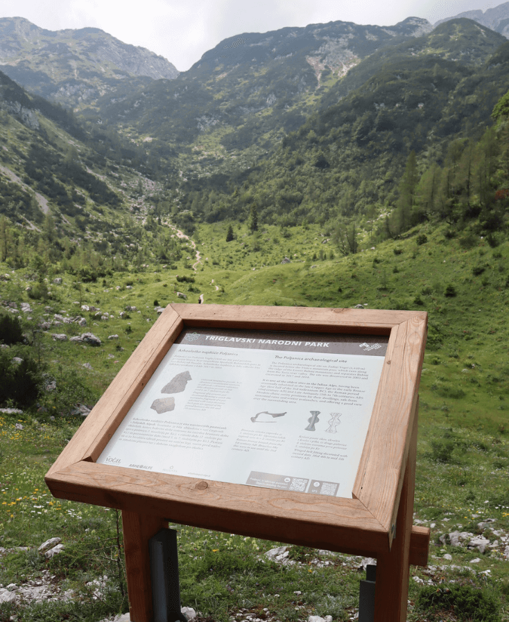 Slikovit pogled na arheološko najdišče na Voglu s skalnatim terenom in informacijsko tablo pod jasnim modrim nebom.